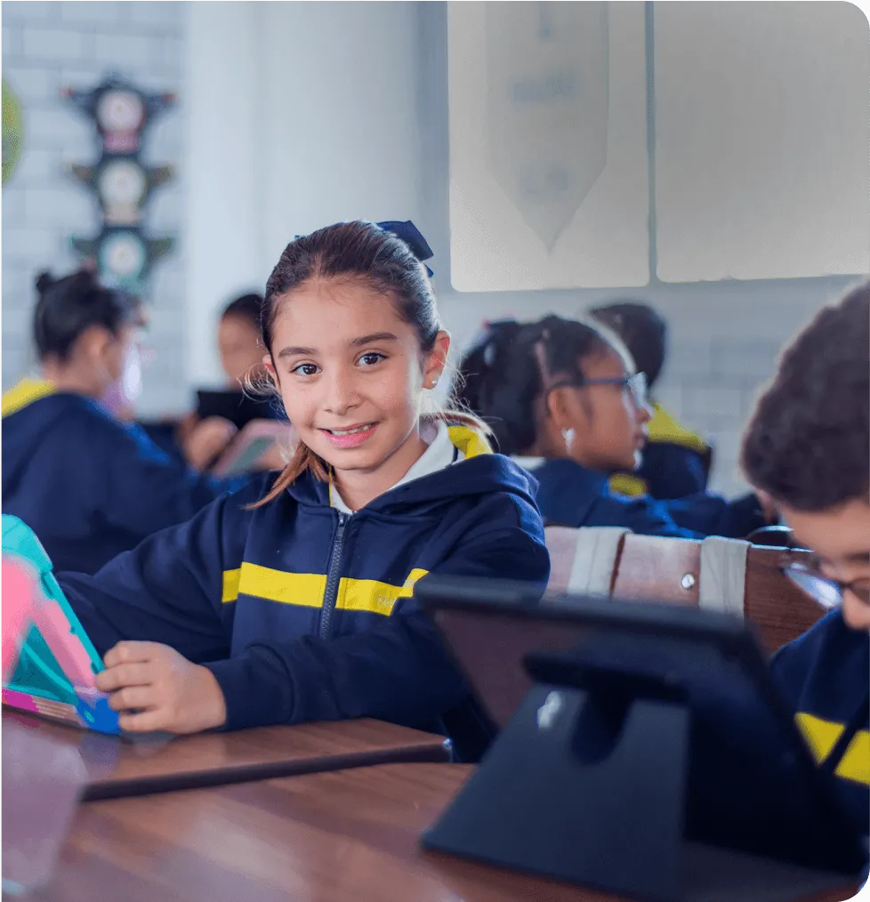 Niña de primaria sonríe frente a su tableta en el salón, con uniforme marino y amarillo