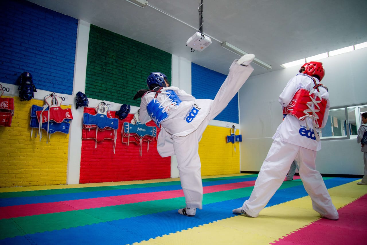 Dos alumnos con uniforme blanco practican taekwondo en el dojo del colegio, con tapetes de colores