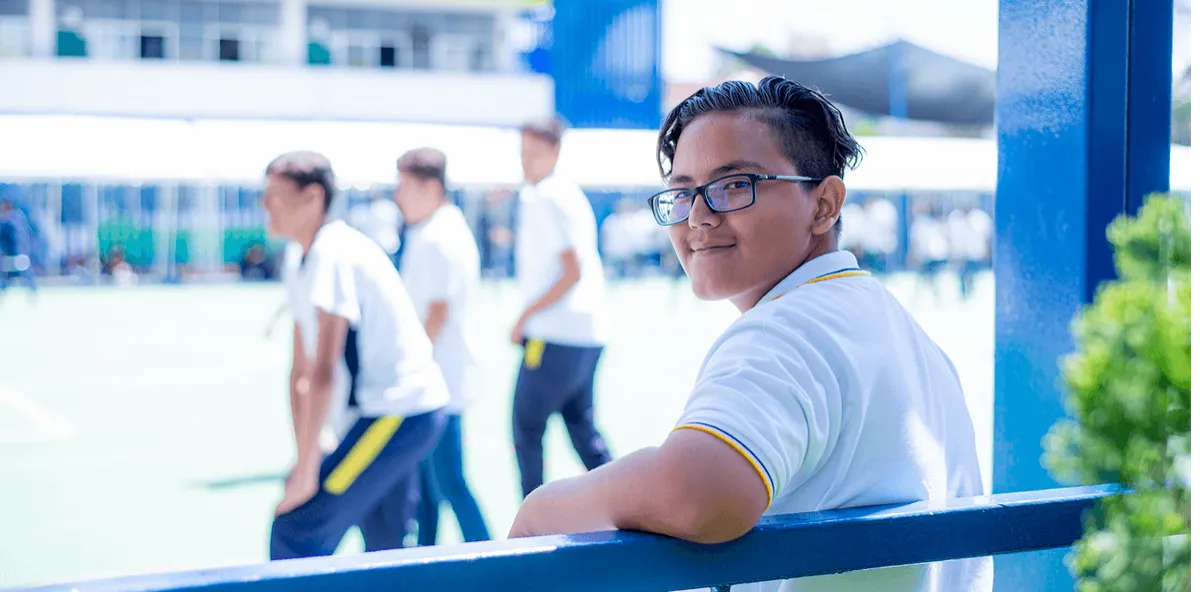 Alumno con playera blanca sentado en una banca azul mientras otros estudiantes caminan en el patio